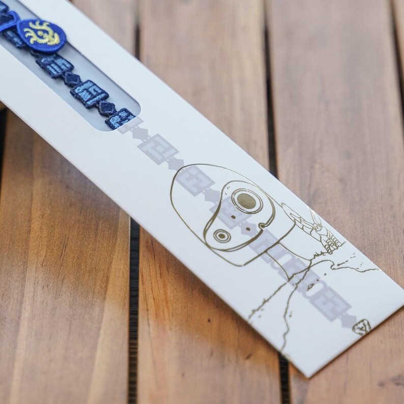 K&ouml;nnte beinhalten: Ein blaues Armband mit aufwendigen Details wird in einer wei&szlig;en Schachtel mit einer Zeichnung eines Roboters pr&auml;sentiert. Die Schachtel ist auf einer Holzoberfl&auml;che schr&auml;g angeordnet. Das Armband hat quadratische und kreisf&ouml;rmige Formen.