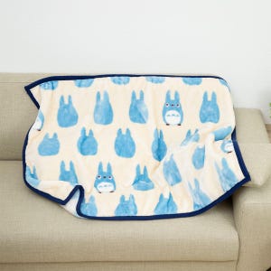 May include: A cream-colored blanket with a repeating pattern of blue cartoon characters. The blanket has a dark blue border and is draped over a beige sofa. The characters are in various poses.