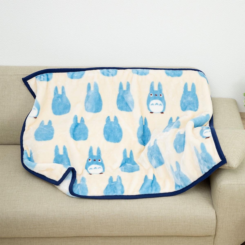 May include: A cream-colored blanket with a repeating pattern of blue cartoon characters. The blanket has a dark blue border and is draped over a beige sofa. The characters are in various poses.