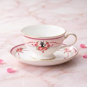 May include: A white teacup and saucer set with a pink floral design and gold trim. The teacup features a black cat illustration surrounded by red roses. The saucer mirrors the teacup's design, creating a cohesive tea set.