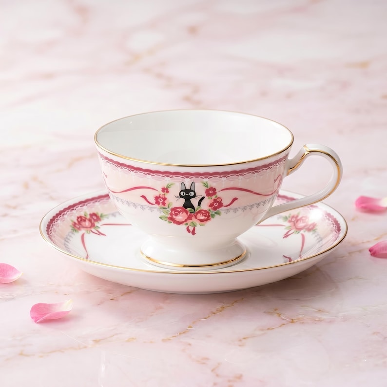 May include: A white teacup and saucer set with a pink floral design and gold trim. The teacup features a black cat illustration surrounded by red roses. The saucer mirrors the teacup's design, creating a cohesive tea set.