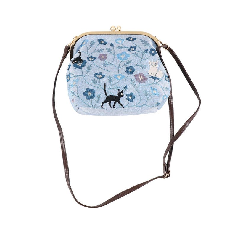 May include: A light blue crossbody bag with a floral and cat embroidery design. The bag features a gold-colored clasp and a brown strap. The embroidered design includes blue, purple, and white flowers, along with a black cat.