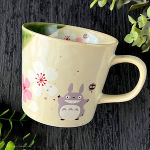 May include: Cream ceramic mug with a cartoon character and floral design. The mug has a curved handle and a green rim. The character is grey with a white belly. Pink and white flowers are scattered around the mug.