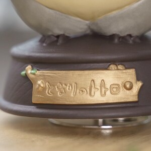 May include: A close-up of a decorative music box base. The base is brown with a gold-colored plaque that has Japanese text. The music box has a light-colored top.