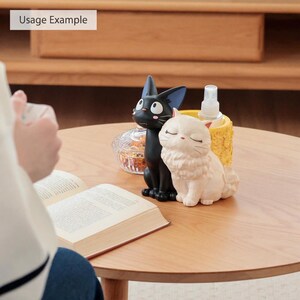 Puede incluir: Una figura de gato negro con ojos grandes y una figura de gato blanco con ojos cerrados, ambas colocadas frente a un soporte cil&iacute;ndrico amarillo texturizado que contiene un atomizador transparente, junto a un peque&ntilde;o recipiente de vidrio.