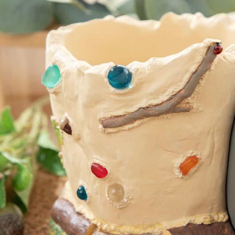 Puede incluir: Una maceta de cer&aacute;mica beige con una superficie texturizada, adornada con coloridos acentos en forma de gema. La maceta presenta un detalle en forma de rama y un interior de color claro, adecuada para plantas peque&ntilde;as o uso decorativo.