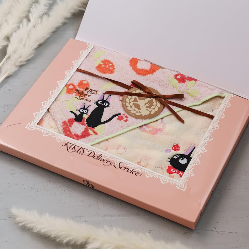 May include: A pink gift box containing a Kiki's Delivery Service themed towel set. The towels display a floral pattern and a black cat illustration. The box is tied with a brown ribbon and has the text "KIKIS Delivery Service".