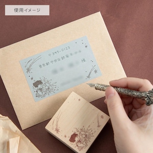 May include: A light brown envelope with a rectangular address label featuring a light blue background, star illustrations, and Japanese text. A square wooden rubber stamp with a matching star and character design sits below the envelope.