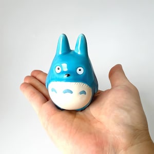 May include: A blue ceramic figurine of a cartoon character with large eyes and pointed ears. The figurine has a white belly and is held in a person's open hand against a white background. The figurine is a popular character from an animated film.