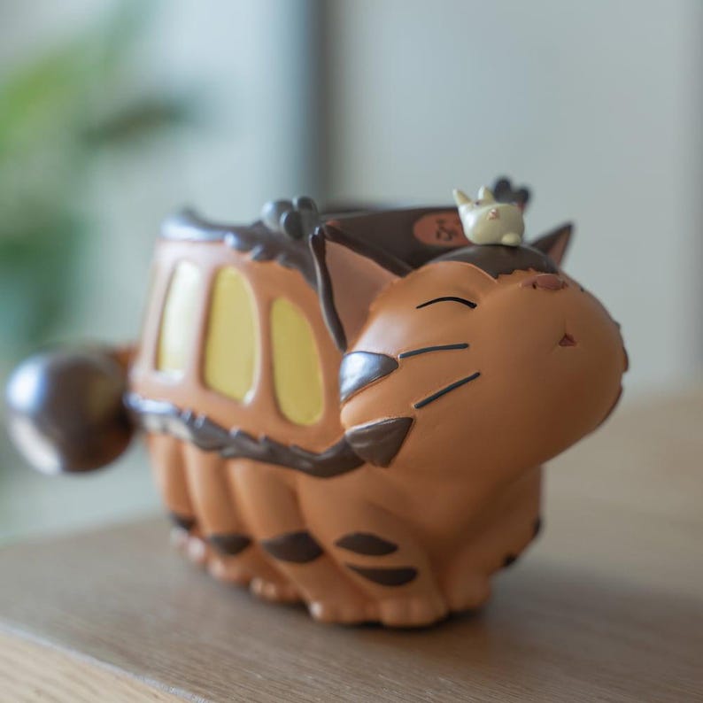 May include: A brown and orange figurine of a cat-like creature with a bus-like body. The figurine has yellow window details and a small white creature on top. The figurine is on a wooden surface.