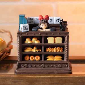 Pode incluir: Uma exibi&ccedil;&atilde;o de padaria em miniatura marrom com uma menina com um la&ccedil;o vermelho, um gato preto e v&aacute;rios produtos de panifica&ccedil;&atilde;o. A exibi&ccedil;&atilde;o inclui um calend&aacute;rio mostrando 20 de fevereiro e um pequeno cubo com um design de bruxa.