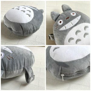 May include: A plush, gray Totoro-shaped pillow with a white belly and embroidered details. The pillow features a zipper closure and a small loop. The design is based on the animated character.