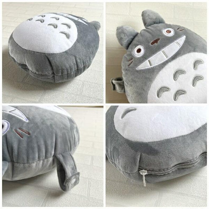 May include: A plush, gray Totoro-shaped pillow with a white belly and embroidered details. The pillow features a zipper closure and a small loop. The design is based on the animated character.