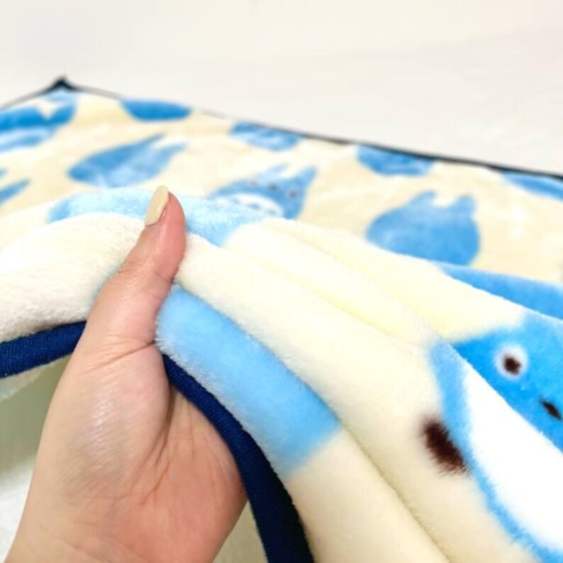 May include: A soft, plush blanket featuring a repeating pattern of light blue cartoon characters on a cream-colored background. The blanket has a light blue border and is folded, showing the soft texture of the fabric.