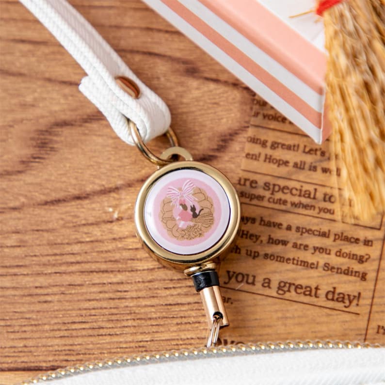 May include: A gold-toned retractable badge holder with a pink and gold design, featuring a white strap. The holder has a circular design with a pink background and a gold floral and silhouette design. The strap is white.