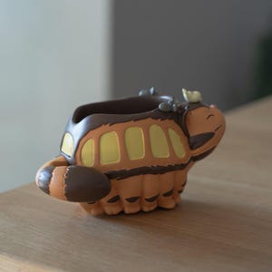 May include: A whimsical, orange and brown planter shaped like a cartoon character. The planter has a bus-like design with yellow window details and a smiling face. The tail and head feature darker brown accents, adding depth to the design.