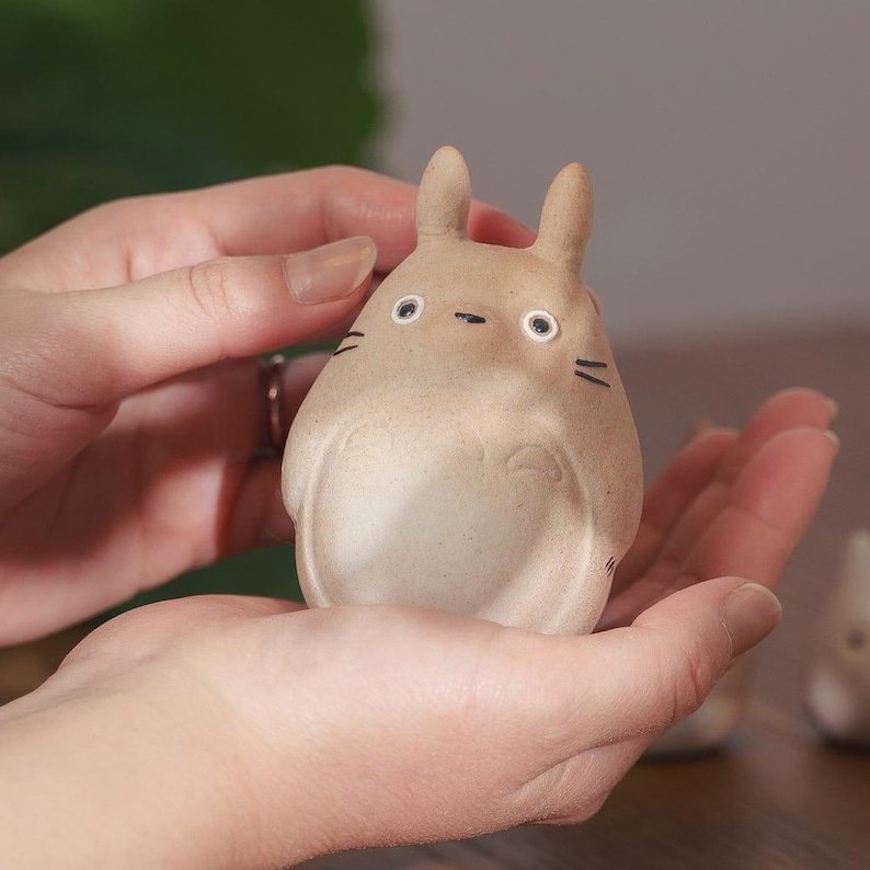 May include: A tan-colored ceramic figurine of a Totoro character, with black facial details and small ears. The figurine is held in open hands, showcasing its rounded body and detailed features. The figurine is a collectible item.