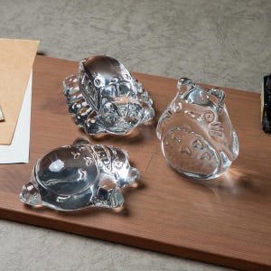 May include: Three clear glass figurines of characters from the animated film "My Neighbor Totoro" are displayed on a wooden surface. The figurines include a bus, a Totoro, and a smaller creature, all with detailed features.