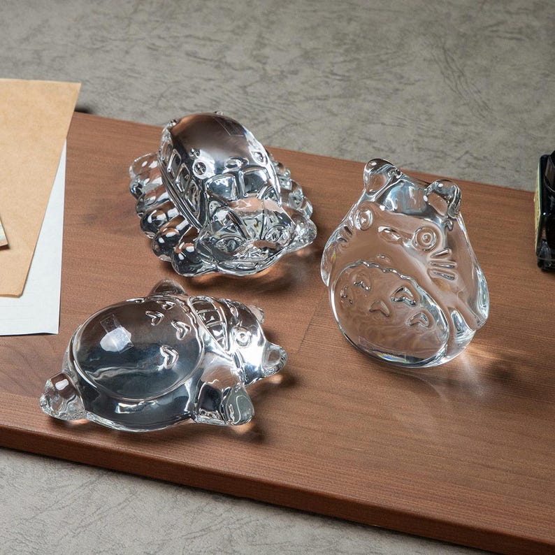 May include: Three clear glass figurines of characters from the animated film "My Neighbor Totoro" are displayed on a wooden surface. The figurines include a bus, a Totoro, and a smaller creature, all with detailed features.