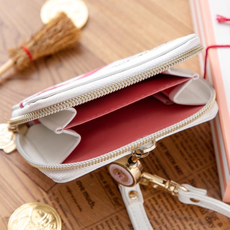 May include: White, zippered coin purse with a gold-colored zipper and accents. The interior is a vibrant pink, with multiple compartments. A small wrist strap and charm are attached. The purse is open, revealing the interior.