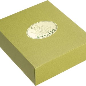 May include: A rectangular, olive-green gift box with a textured surface. An oval-shaped, gold-colored emblem is affixed to the top, featuring a cartoon character and Japanese text. The box is closed and appears to be empty.