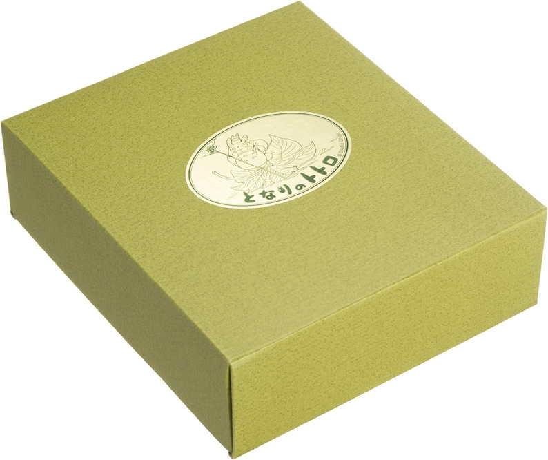 May include: A rectangular, olive-green gift box with a textured surface. An oval-shaped, gold-colored emblem is affixed to the top, featuring a cartoon character and Japanese text. The box is closed and appears to be empty.