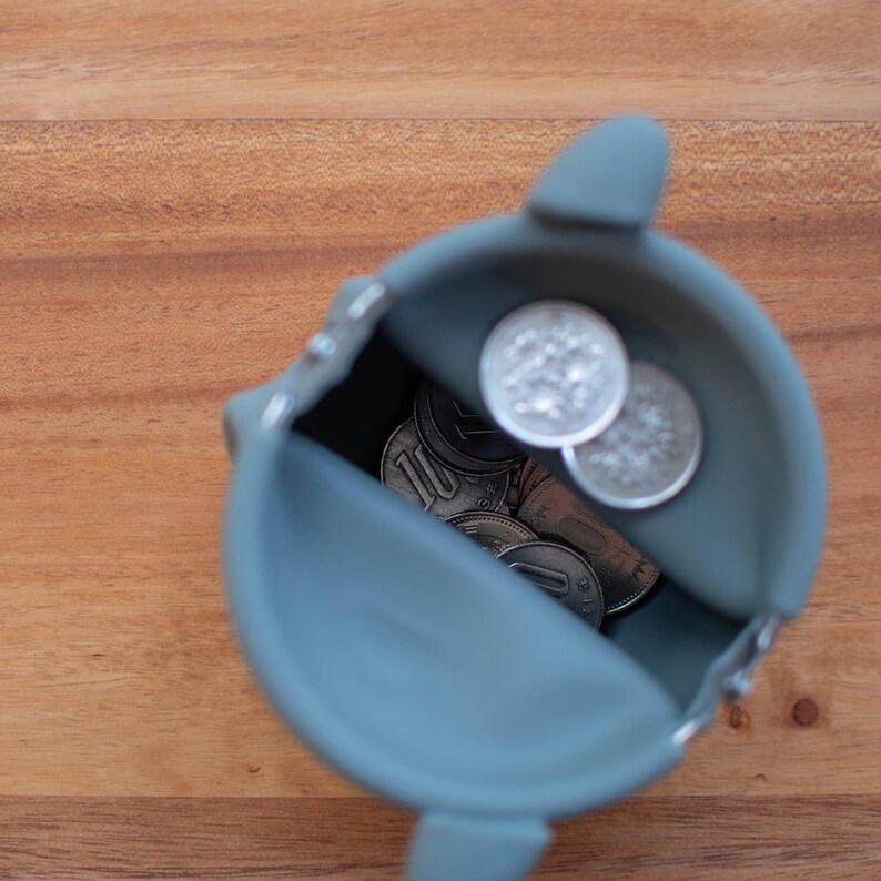 May include: A teal-colored coin purse filled with various coins. The purse is open, revealing the contents, and sits on a wooden surface. The purse has a simple, rounded design.