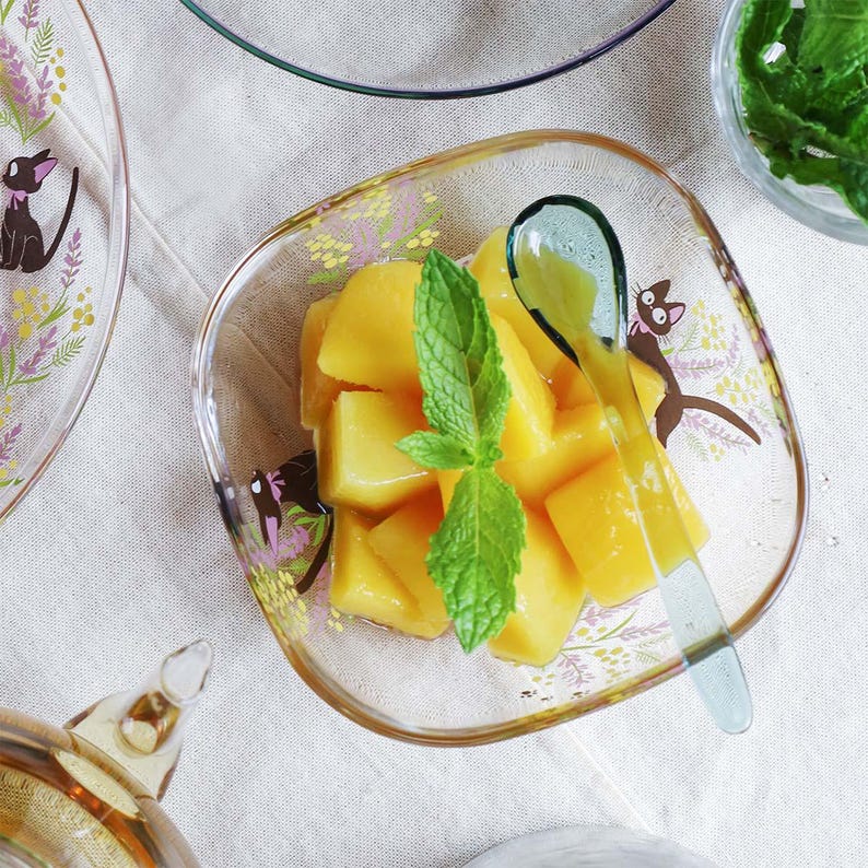 May include: A square glass bowl with rounded corners featuring a black cat illustration and floral patterns, filled with cubed yellow mango pieces topped with a fresh green mint sprig and a translucent green spoon resting inside.