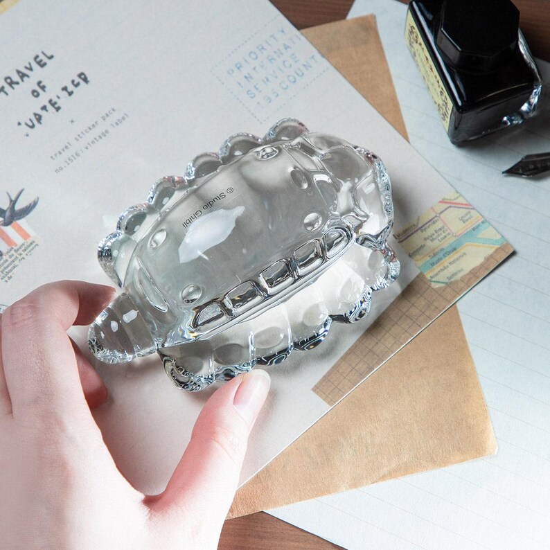 May include: A clear glass ashtray with a scalloped edge and a rectangular design. The ashtray is sitting on top of a vintage paper with the text "TRAVEL OF CUPTE' IER". A bottle of black ink and a pen are also visible.