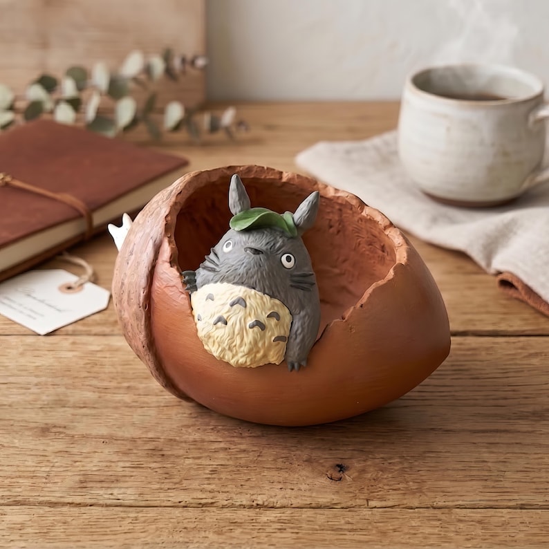 K&ouml;nnte beinhalten: Ein dekorativer, brauner, eichelf&ouml;rmiger Beh&auml;lter mit einer grauen Totoro-Figur im Inneren. Der Totoro hat ein gr&uuml;nes Blatt auf dem Kopf und einen cremefarbenen Bauch. Ein braunes Notizbuch und eine wei&szlig;e Tasse sind im Hintergrund.