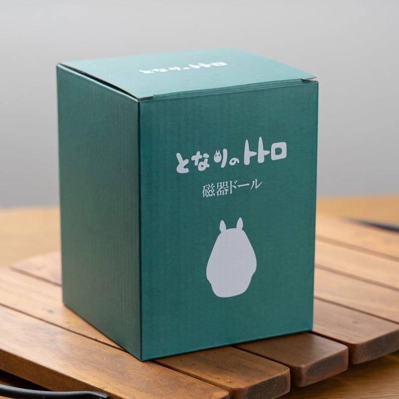 Puede incluir: Una caja verde azulado con texto japon&eacute;s y una silueta blanca de Totoro. La caja est&aacute; sobre una superficie de madera. El texto en la caja dice "となりのトトロ" y "磁器ドール".