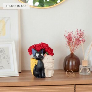 Puede incluir: Una maceta decorativa con un gato negro y un gato blanco sentados uno al lado del otro, sobre una base amarilla texturizada. La maceta est&aacute; llena de claveles rojos brillantes, creando un arreglo colorido sobre una superficie de madera.