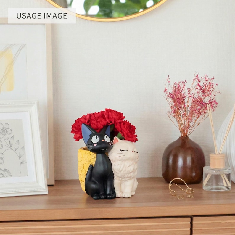 Puede incluir: Una maceta decorativa con un gato negro y un gato blanco sentados uno al lado del otro, sobre una base amarilla texturizada. La maceta est&aacute; llena de claveles rojos brillantes, creando un arreglo colorido sobre una superficie de madera.