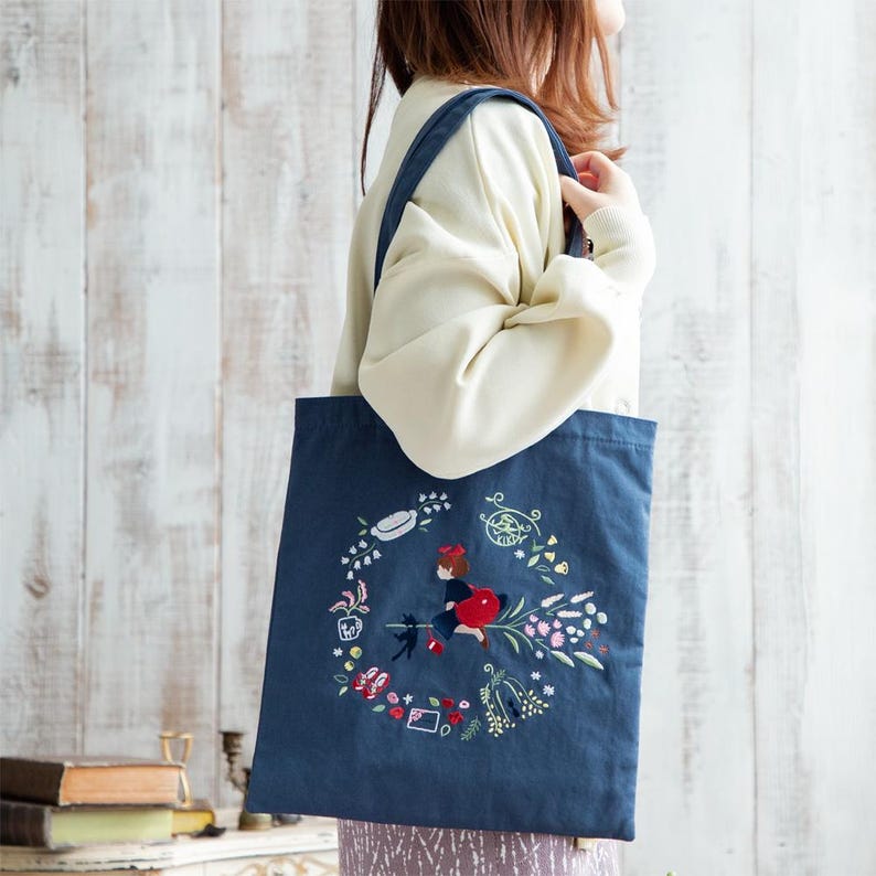 K&ouml;nnte beinhalten: Eine blaue Canvas-Tragetasche mit einem gestickten Design, das ein M&auml;dchen zeigt, das auf einer schwarzen Katze mit einer roten Tasche reitet. Das Design ist von Blumen und anderen kleinen Details umgeben.