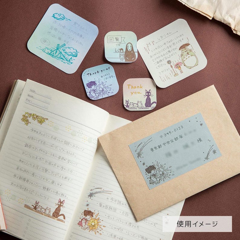 May include: An open notebook with lined pages featuring handwritten Japanese text and small illustrations of cats, alongside a brown envelope with a blue address label. Five square and rectangular stickers with various anime-style illustrations are included.