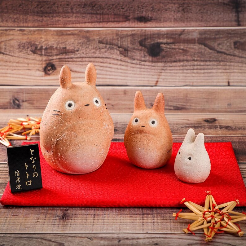 May include: Three ceramic Totoro figurines in varying sizes, with tan and white coloring, sit on a red mat. The largest has a detailed face, while the smallest is white. A small black sign with Japanese text is on the left.