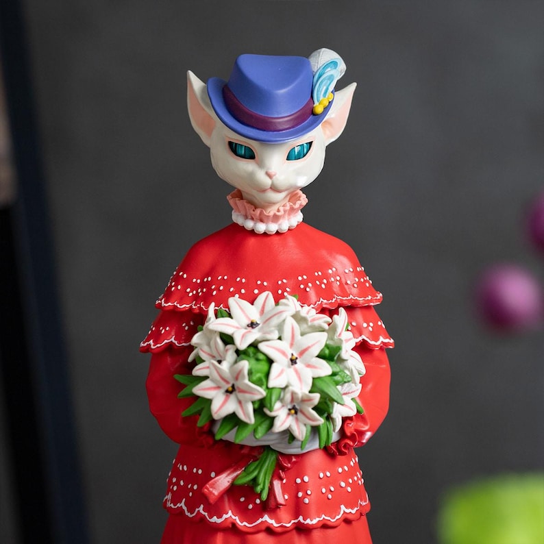 Puede incluir: Una figura caprichosa de un personaje felino con un vestido rojo y un sombrero azul con una pluma. La figura sostiene un ramo de flores blancas. El gato tiene ojos azules y un cuello con volantes rosas. Un art&iacute;culo decorativo &uacute;nico.