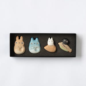 Puede incluir: Un juego de cuatro reposa palillos de cer&aacute;mica dispuestos en una caja rectangular negra. Las figuras incluyen una criatura color canela, una azul, una blanca y un duende del holl&iacute;n negro sobre una hoja verde con texto japon&eacute;s.