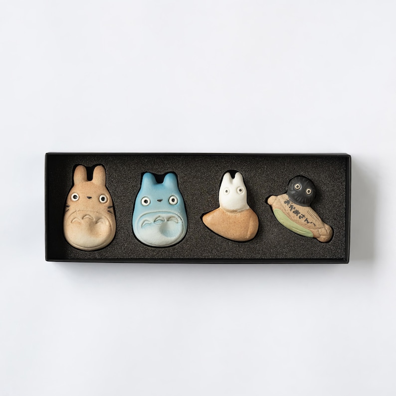 Puede incluir: Un juego de cuatro reposa palillos de cer&aacute;mica dispuestos en una caja rectangular negra. Las figuras incluyen una criatura color canela, una azul, una blanca y un duende del holl&iacute;n negro sobre una hoja verde con texto japon&eacute;s.