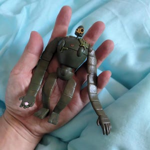 Peut inclure: Figurine de robot vert olive avec des membres articul&eacute;s et une petite t&ecirc;te en forme de cr&acirc;ne. Le robot a une apparence us&eacute;e, avec des articulations visibles et des d&eacute;tails ressemblant &agrave; de la mousse. La figurine est tenue dans une main sur un fond bleu clair.