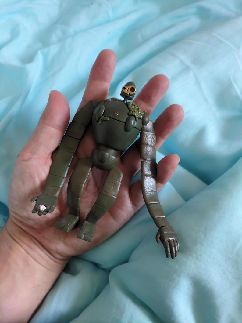 Peut inclure: Figurine de robot vert olive avec des membres articul&eacute;s et une petite t&ecirc;te en forme de cr&acirc;ne. Le robot a une apparence us&eacute;e, avec des articulations visibles et des d&eacute;tails ressemblant &agrave; de la mousse. La figurine est tenue dans une main sur un fond bleu clair.