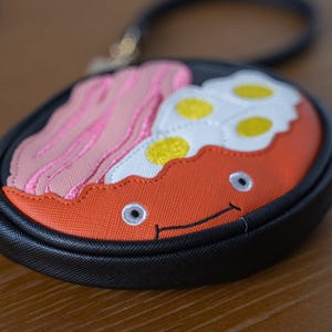 May include: A novelty round coin purse with a black handle. The purse features an embroidered design of a breakfast plate with bacon, fried eggs, and a smiling face. The background is a blurred wooden surface.