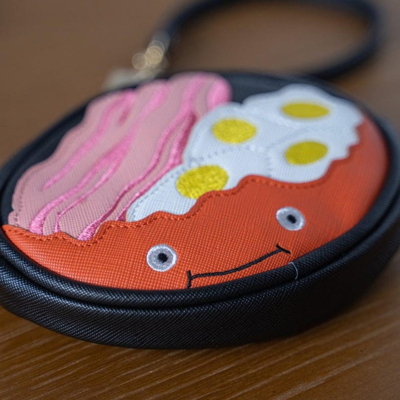 May include: A novelty round coin purse with a black handle. The purse features an embroidered design of a breakfast plate with bacon, fried eggs, and a smiling face. The background is a blurred wooden surface.