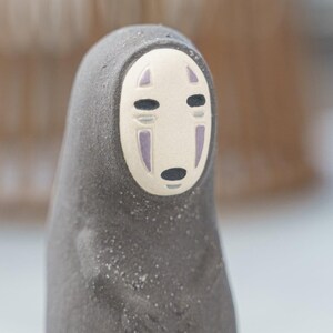 Puede incluir: Una peque&ntilde;a figura de color gris oscuro con una superficie texturizada y mate. El personaje presenta una m&aacute;scara facial ovalada de color crema lisa con dos ojos horizontales negros, una peque&ntilde;a boca ovalada negra y dos marcas verticales moradas.