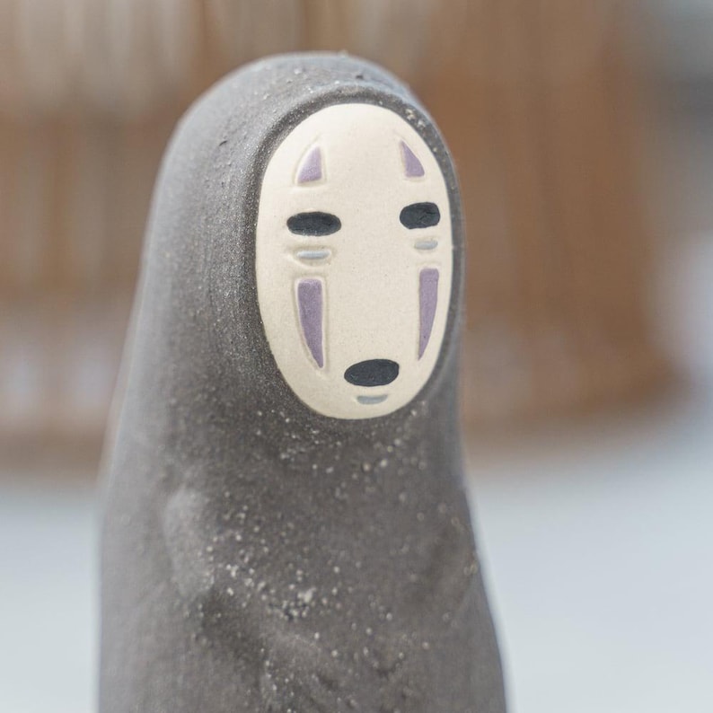 Puede incluir: Una peque&ntilde;a figura de color gris oscuro con una superficie texturizada y mate. El personaje presenta una m&aacute;scara facial ovalada de color crema lisa con dos ojos horizontales negros, una peque&ntilde;a boca ovalada negra y dos marcas verticales moradas.