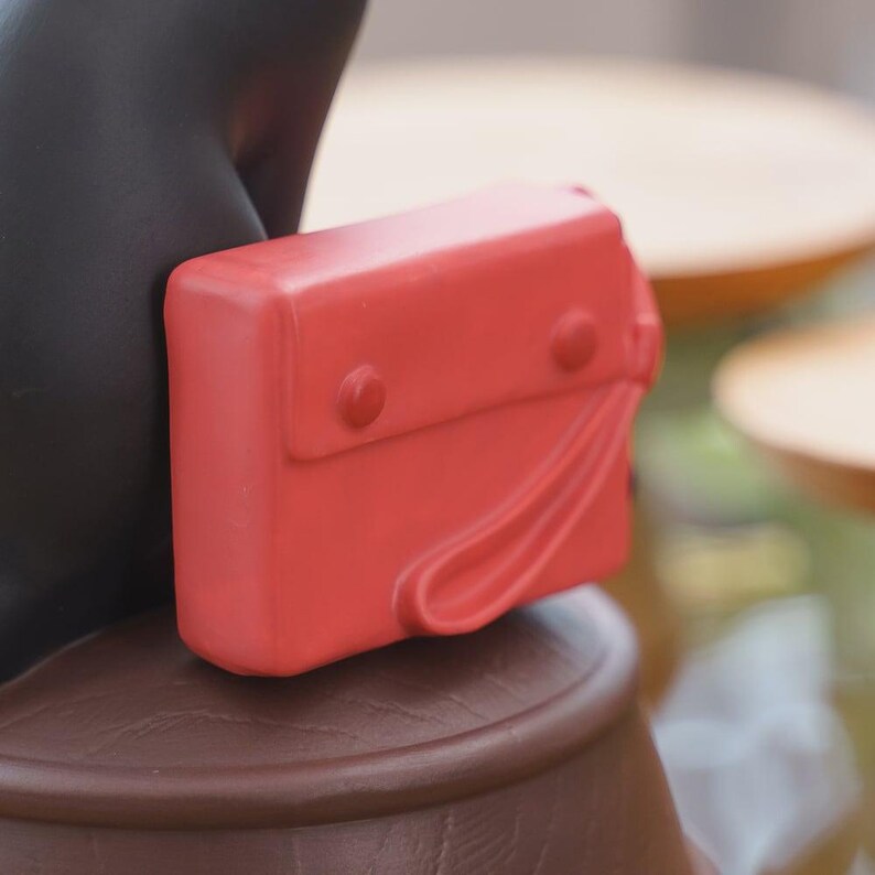 May include: A small, bright red purse with a flap closure and two round button details. The purse has a curved handle and is resting on a brown surface. The background is blurred, with hints of other objects.