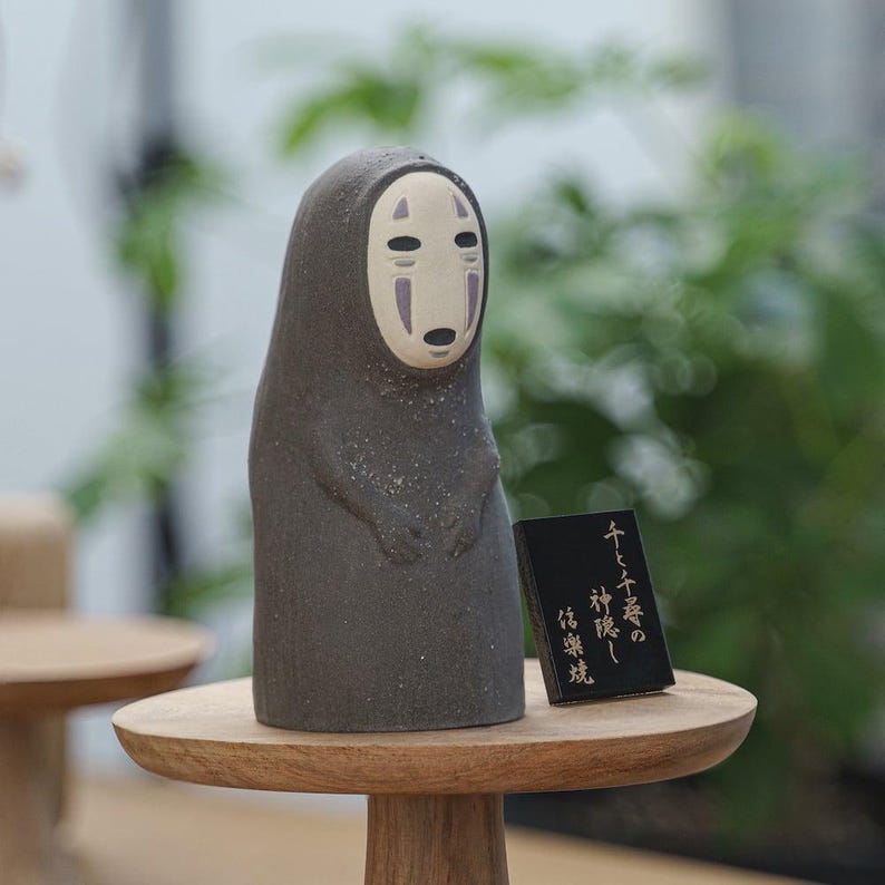 Puede incluir: Una figura de cer&aacute;mica gris oscuro de un personaje con un rostro ovalado, liso y p&aacute;lido, que presenta dos marcas negras en los ojos y una peque&ntilde;a boca negra. Junto a la figura hay un peque&ntilde;o letrero rectangular negro con texto japon&eacute;s en blanco.