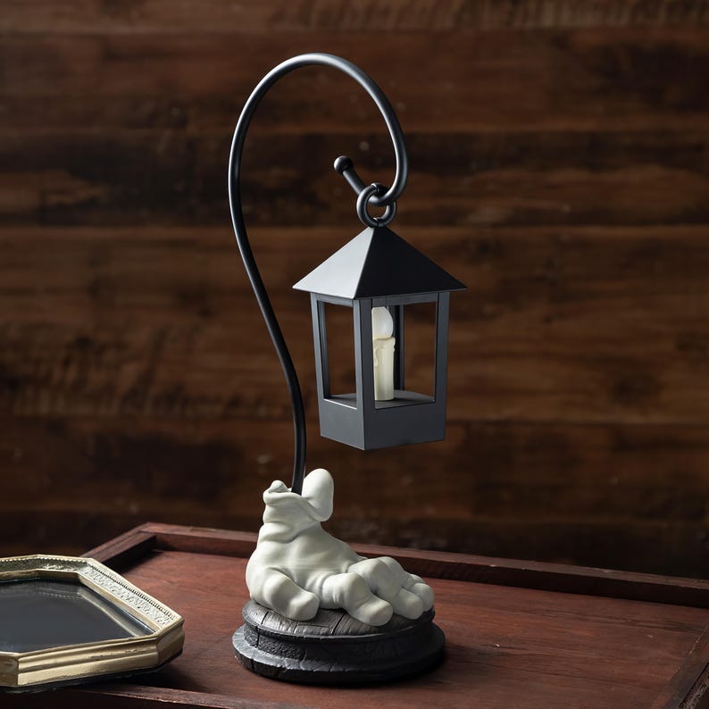 May include: A black metal lantern with a curved support, featuring a small, square lantern with a candle inside. The base is a sculpted figure in a relaxed pose, resting on a dark, textured platform. The background is a wooden surface.