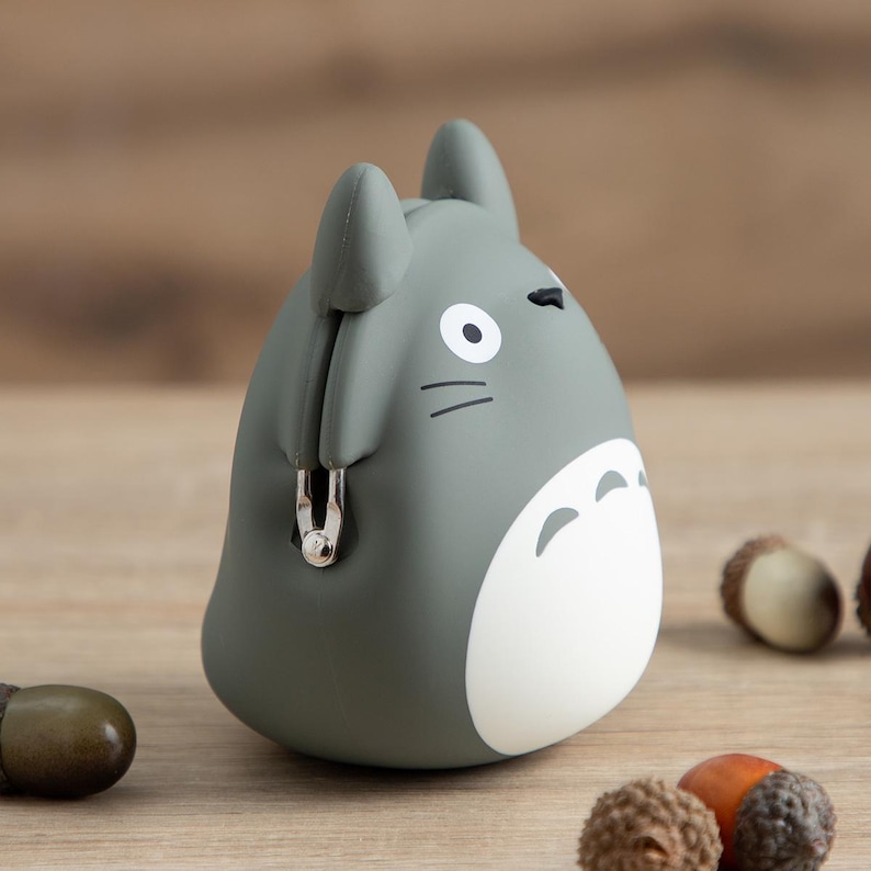 May include: A gray Totoro-shaped coin purse with a white belly and black details. The purse has a metal clasp and two small ears. The background includes acorns.