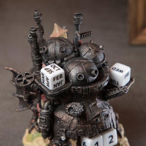 May include: A detailed steampunk-style perpetual calendar sculpture in shades of gray and brown. The sculpture features multiple dice displaying months and days, along with numbered dice. The design includes mechanical-looking components and a small, tan-colored structure.